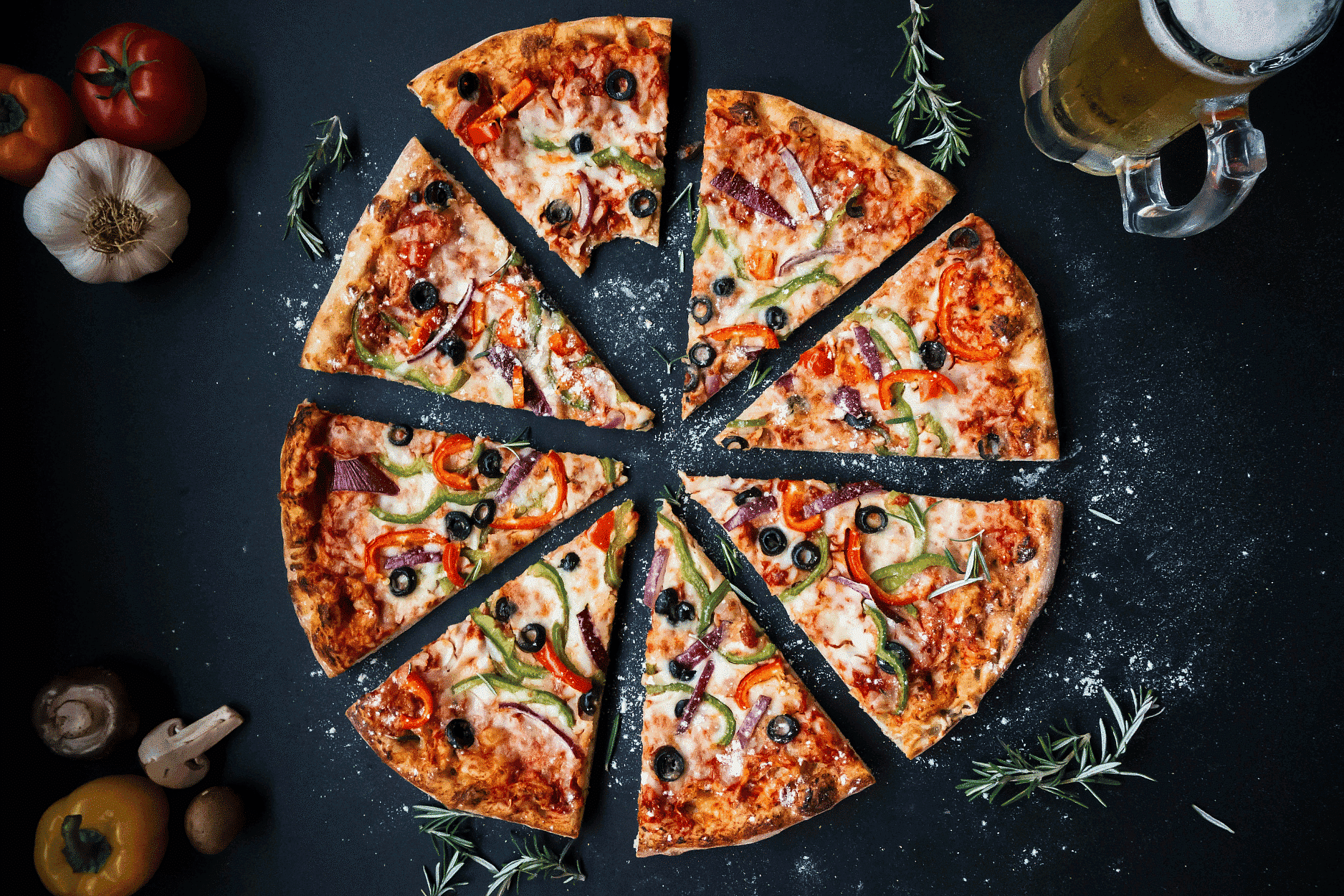 Pizza with various toppings sliced into eight pieces beside a mug of beer and fresh vegetables.